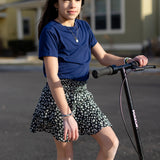 A little girl is wearing a basic navy t-shirt and a black and white skirt. She is standing on a purple and black skooter.