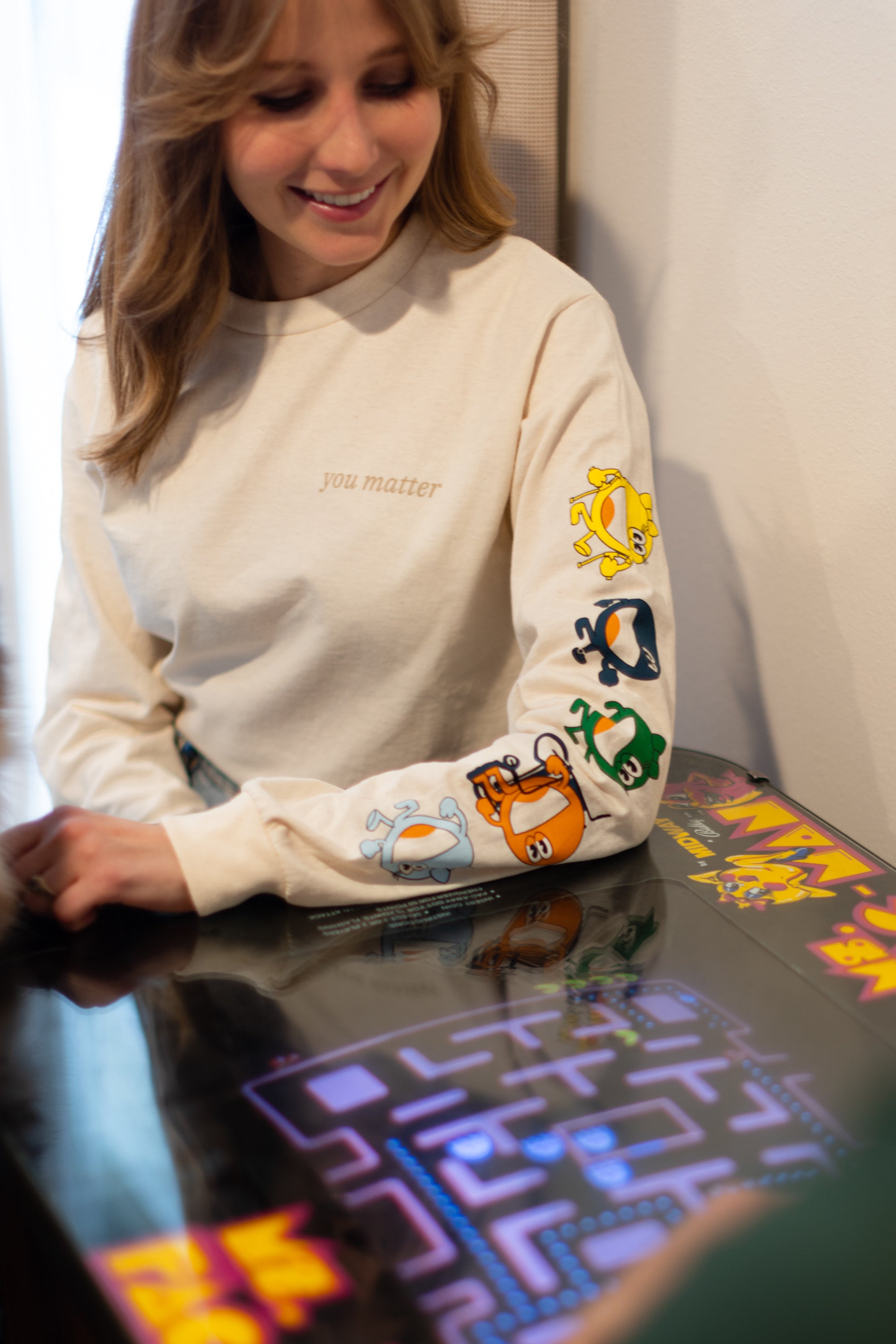 A blonde hair women wearing a cream long sleeve shirt. The cream long sleeve reads 'you matter' on the standard pocket area of the t-shirt. Running down the sleeve is multiple characters: A yellow one, navy, green, red, and light blue. The woman is sitting down playing a vintage game. 