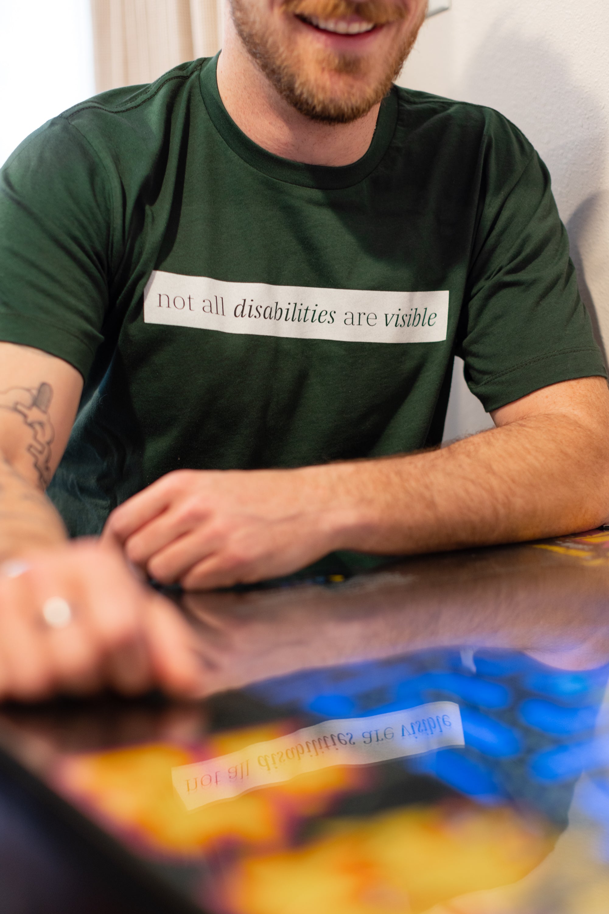 A white man (with a brown beard) wears a forest green short sleeve tee has a beige block printed on the front chest. The words "Not All Disabilities are Visible" are on this block. He is sitting and leans on a table.