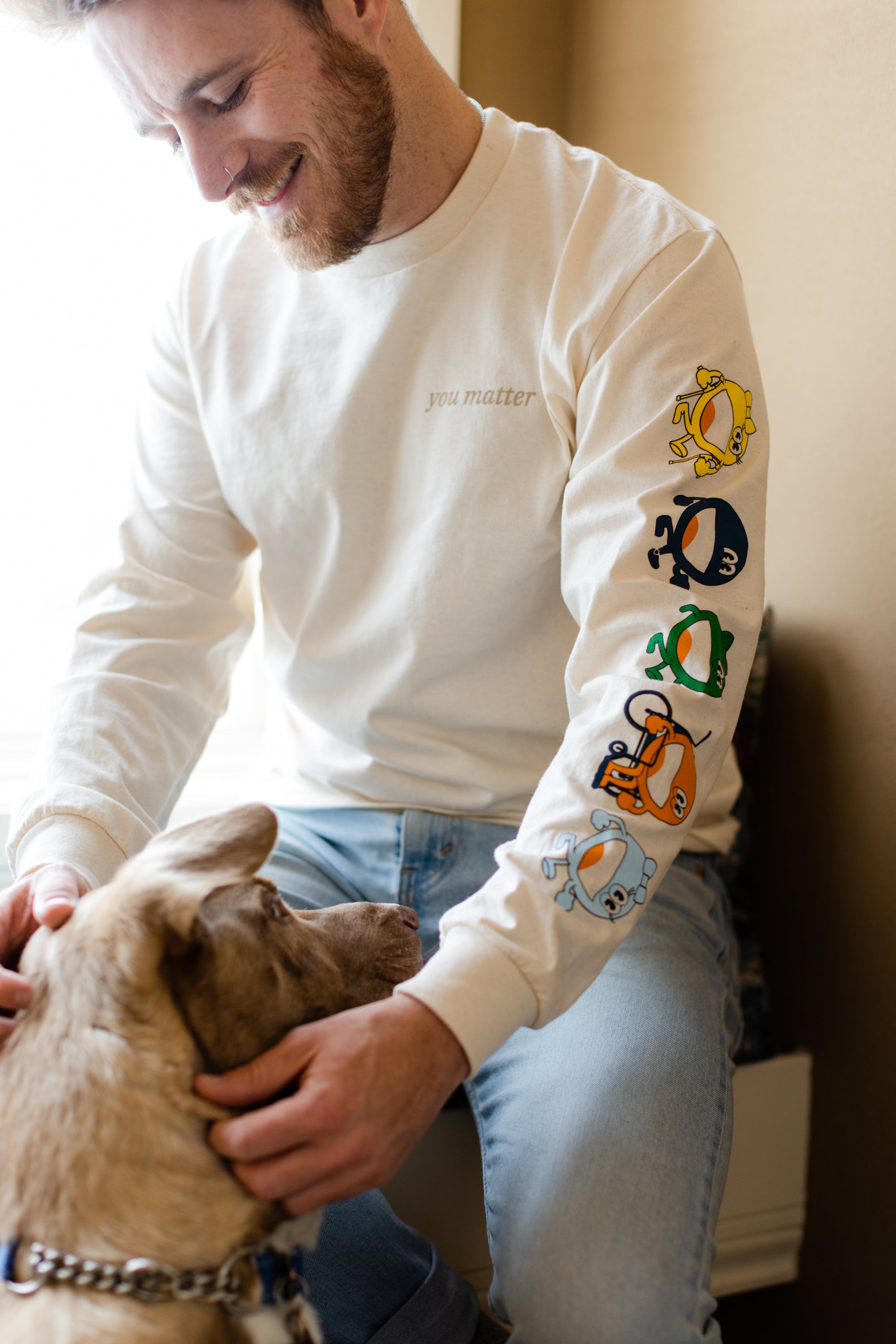 A man is sitting down by a window petting a dog. He is wearing a cream long sleeve shirt that reads 'you matter' on a t-shirt standard pocket area. Running down the sleeve are characters walking down, they are a circle shape. The colors of the characters are yellow, navy, green, orange, and light blue.