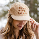 A white woman with long brown hair wears a cream baseball cap with brown text that says "disability is not a bad word."