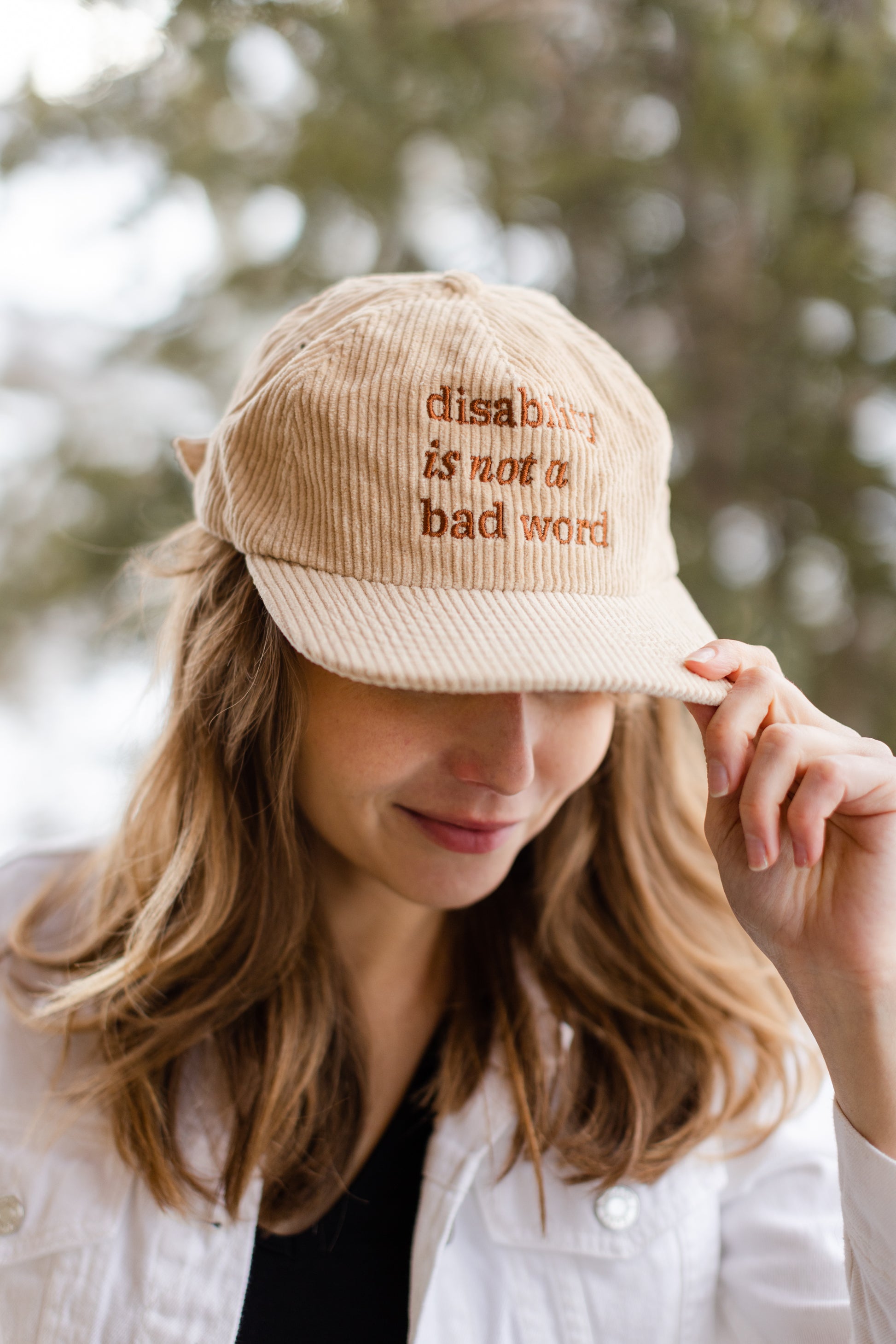 A white woman with long brown hair wears a cream baseball cap with brown text that says "disability is not a bad word."