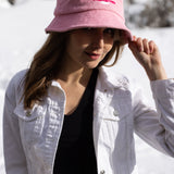 A white woman with brown long hair wears a light pink bucket hat with a white illustration of the sun and pink text that says "Unlimbited." She's in a snowy forest.