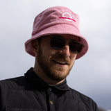 A white man with a beard and brown sunglasses wears a light pink bucket hat with a white illustration of the sun and pink text that says "Unlimbited." He's in a snowy forest.