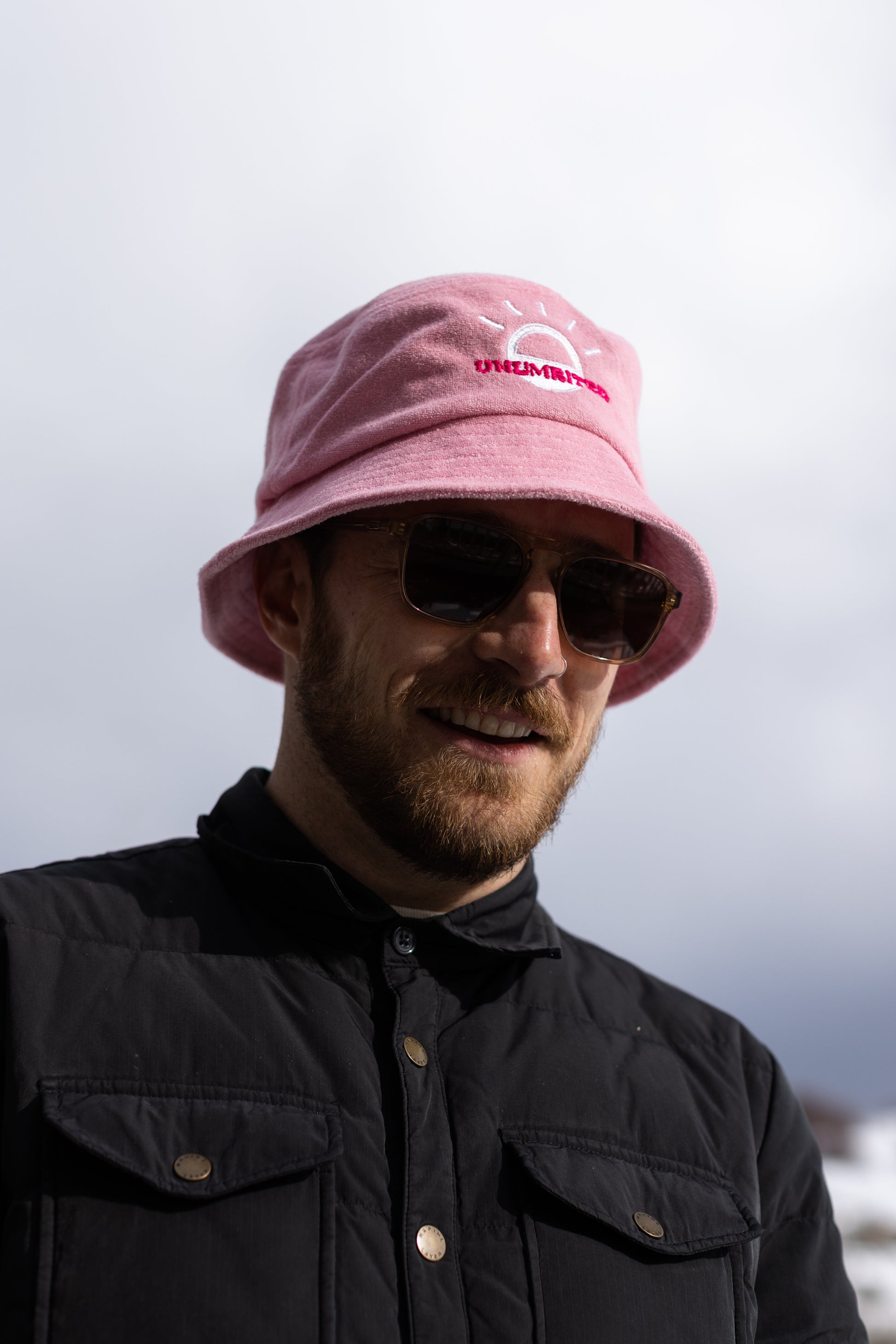 A white man with a beard and brown sunglasses wears a light pink bucket hat with a white illustration of the sun and pink text that says "Unlimbited." He's in a snowy forest.