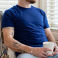 A white man with short brown hair wears a navy t-shirt. He sits on a window seat, holding a mug of coffee.