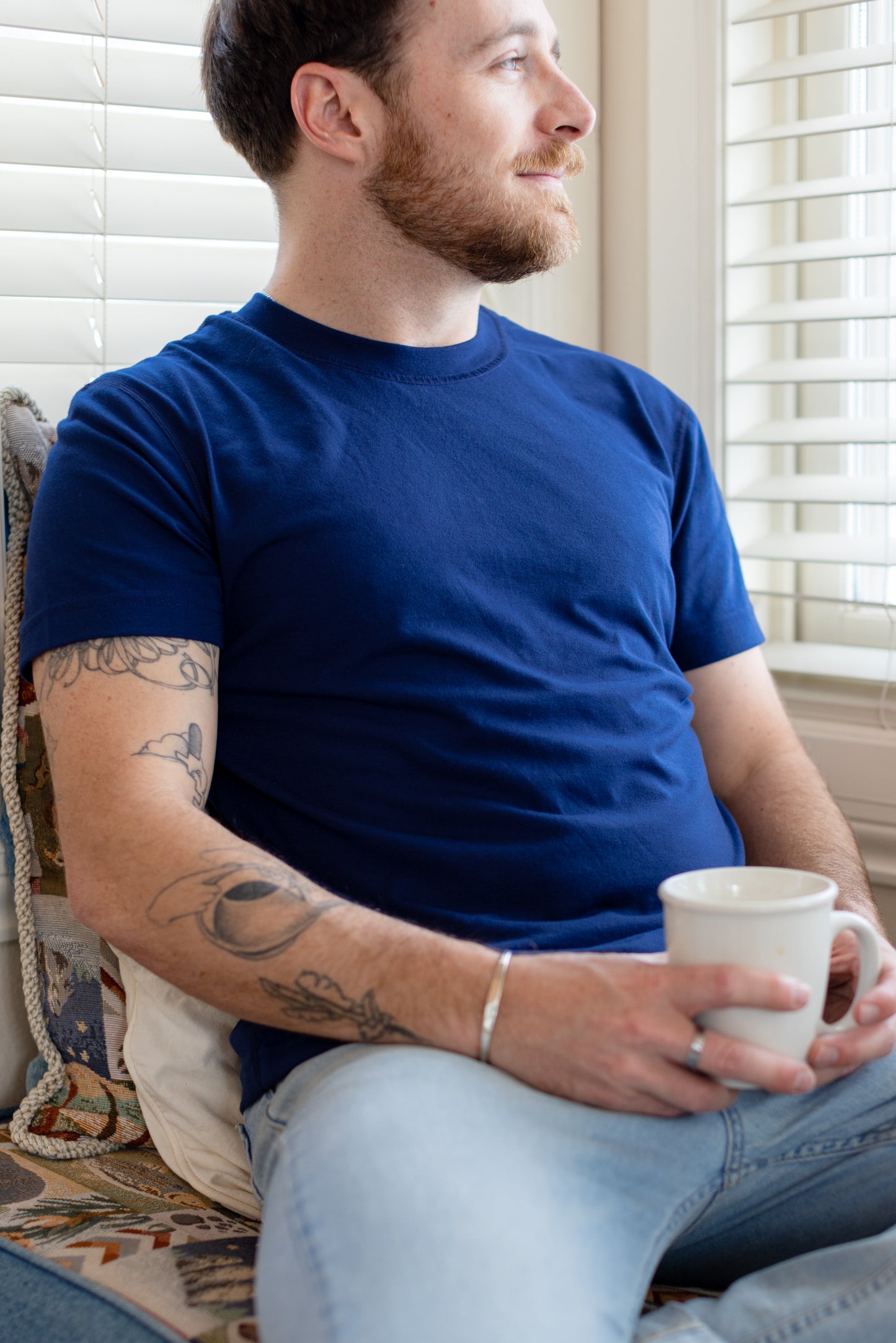 A white man with short brown hair wears a navy t-shirt. He sits on a window seat, holding a mug of coffee.