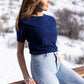 A white woman with long blonde hair is wearing the No Limbits Adaptive Women's Navy Sensory Tee. She leans on a balcony. A snowy hill is in the background.