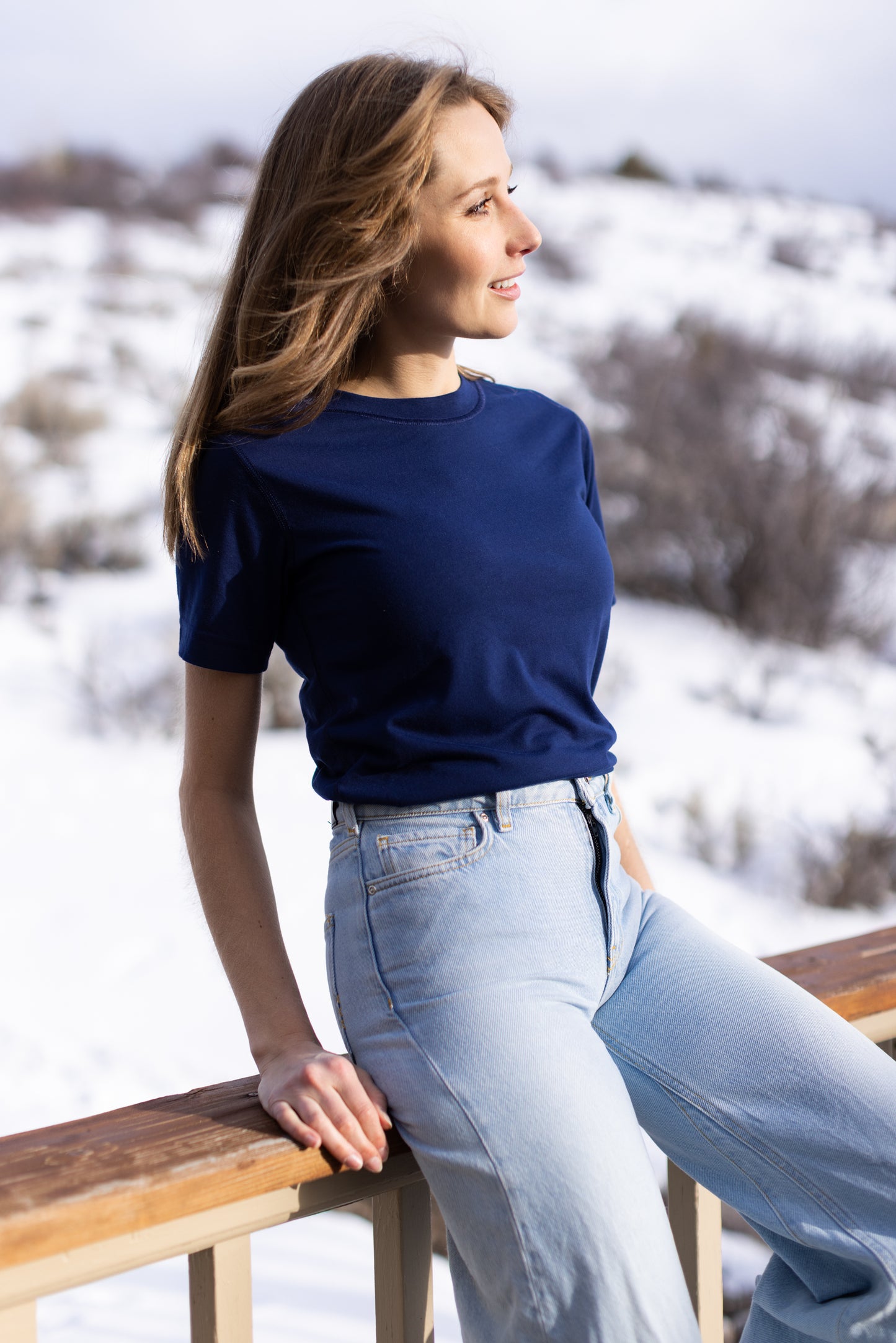 A white woman with long blonde hair is wearing the No Limbits Adaptive Women's Navy Sensory Tee. She leans on a balcony. A snowy hill is in the background.