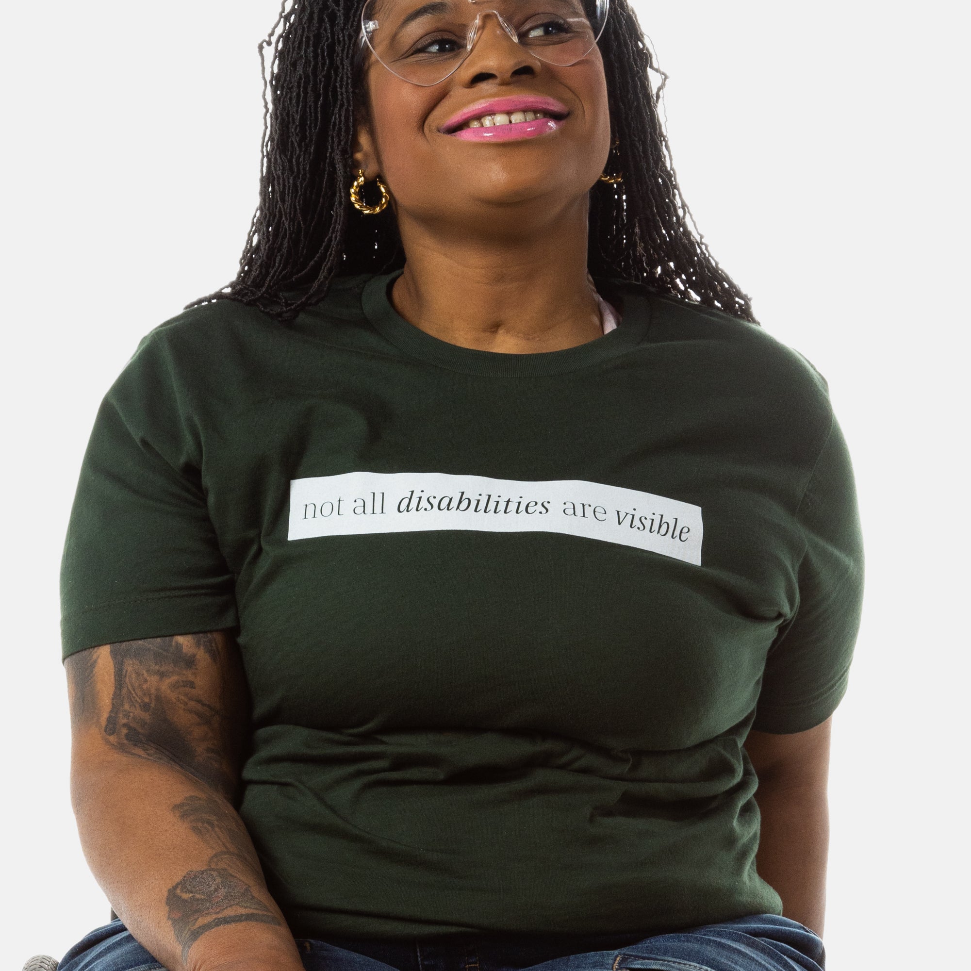 A Black woman (with long black locs, pink lipstick, hoop earrings) wears a forest green short sleeve tee has a beige block printed on the front chest. The words "Not All Disabilities are Visible" are on this block. 