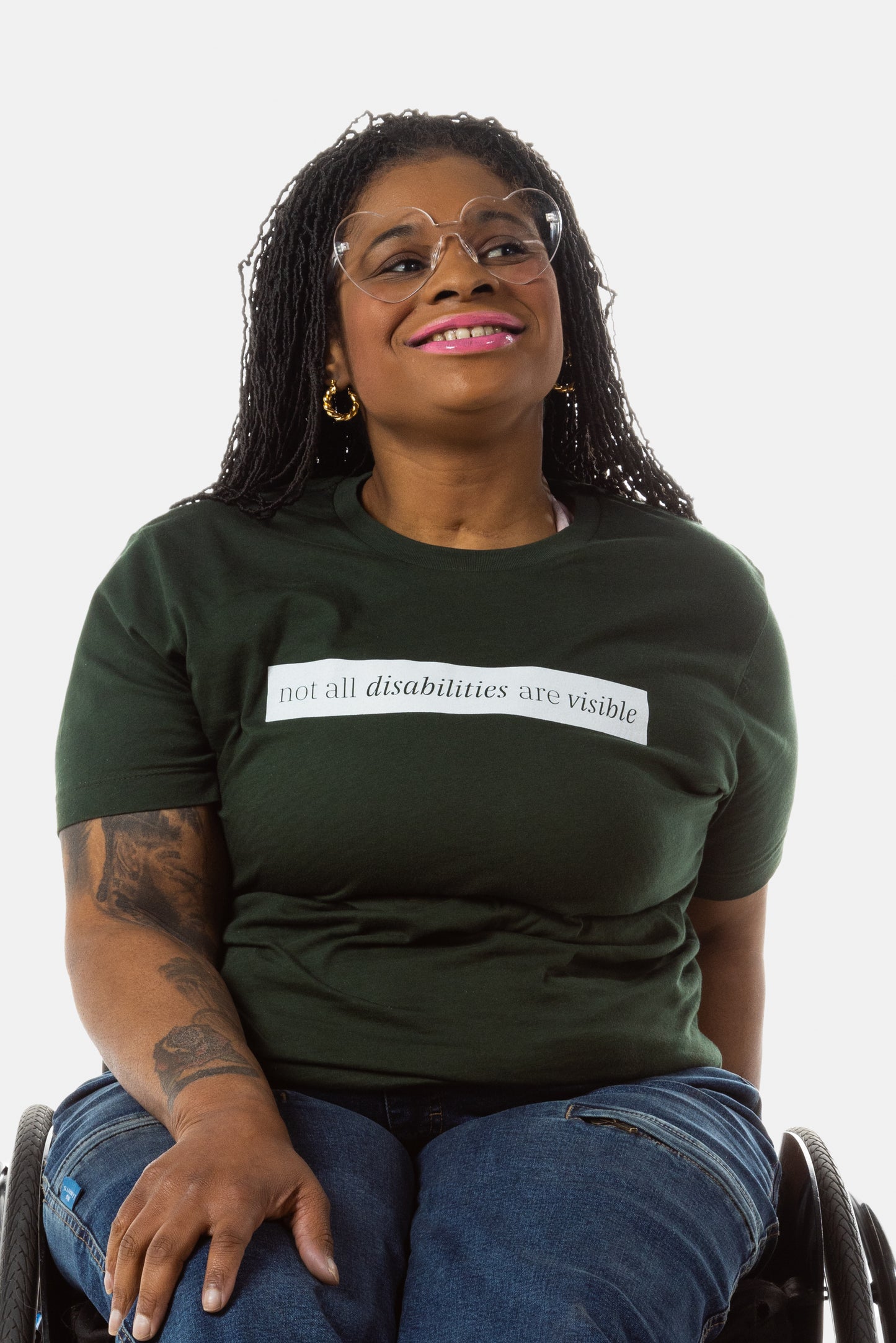 A Black woman (with long black locs, pink lipstick, hoop earrings) wears a forest green short sleeve tee has a beige block printed on the front chest. The words "Not All Disabilities are Visible" are on this block. 