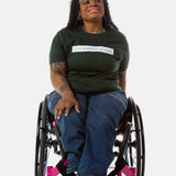 Princess, a Black woman on a pink wheelchair (with long locs, light pink lipstick, clear black sunglasses, and hoops) wears the No Limbits Adaptive Women's Dark Wash Wheelchair Pants.
