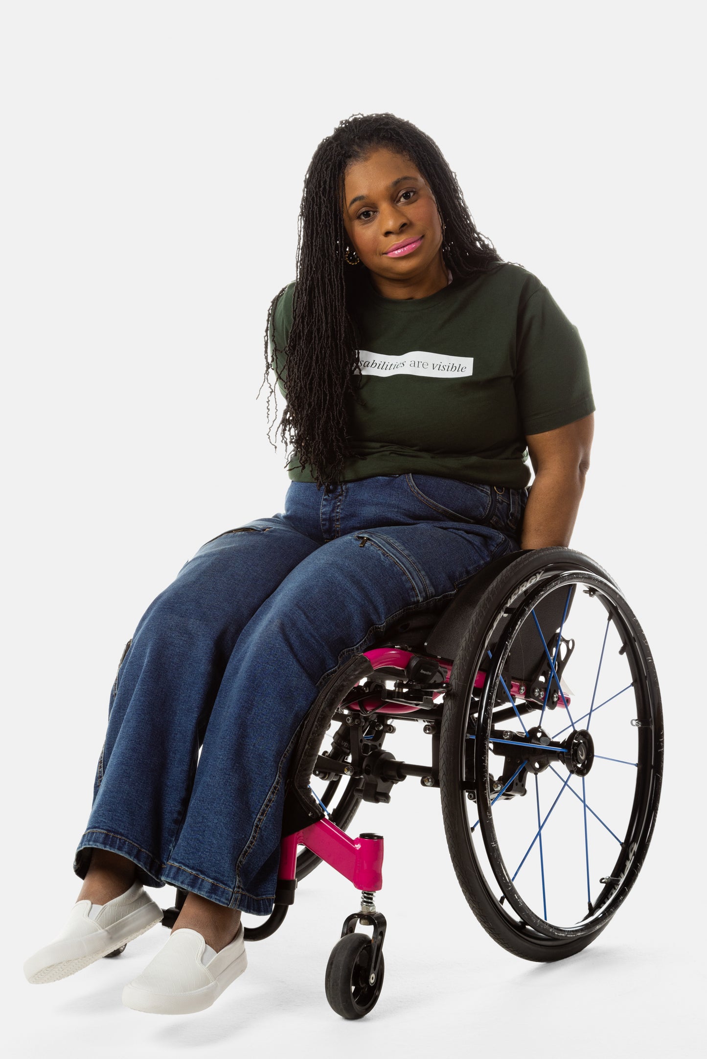 Princess, a Black woman on a pink wheelchair (with long locs, light pink lipstick, clear black sunglasses, and hoops) wears the No Limbits Adaptive Women's Dark Wash Wheelchair Pants.