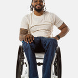 Jessi, a Black man on a white wheelchair (with short locs with a bun on the top of his hair, a goatee, cross earrings, and white sneakers) wears the No Limbits Adaptive Men's Dark Wash Wheelchair Pants.
