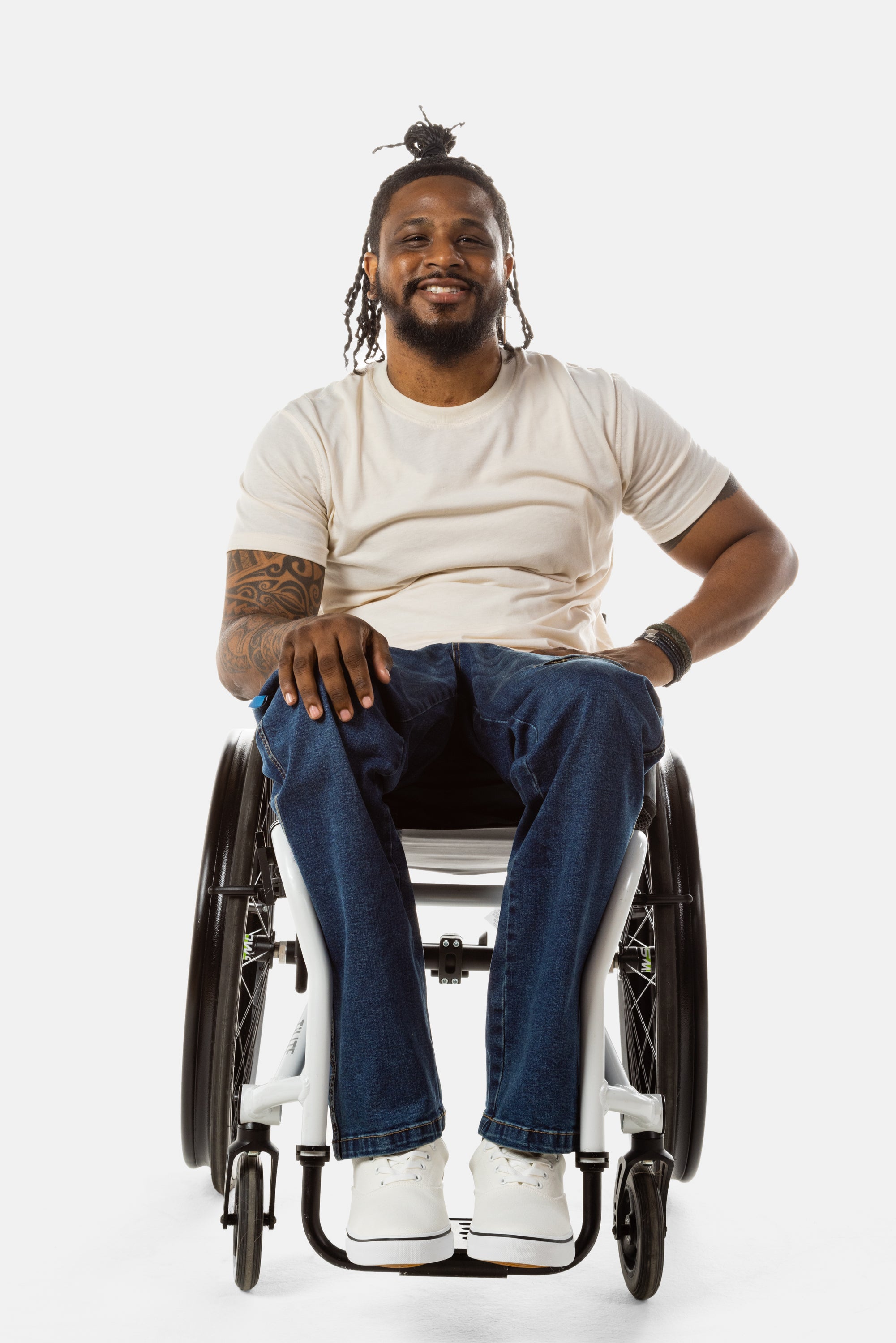 Jessi, a Black man on a white wheelchair (with short locs with a bun on the top of his hair, a goatee, cross earrings, and white sneakers) wears the No Limbits Adaptive Men's Dark Wash Wheelchair Pants.