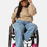 Princess, a Black woman on a pink wheelchair (with long locs, light pink lipstick, clear black sunglasses, and hoops) wears the No Limbits Adaptive Women's Light Wash Wheelchair Pants.