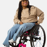 Princess, a Black woman on a pink wheelchair (with long locs, light pink lipstick, clear black sunglasses, and hoops) wears the No Limbits Adaptive Women's Light Wash Wheelchair Pants.
