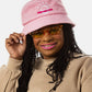 A black woman with sunglasses, pink lipstick, and long black locs wears a light pink bucket hat with a white illustration of the sun and pink text that says "Unlimbited"