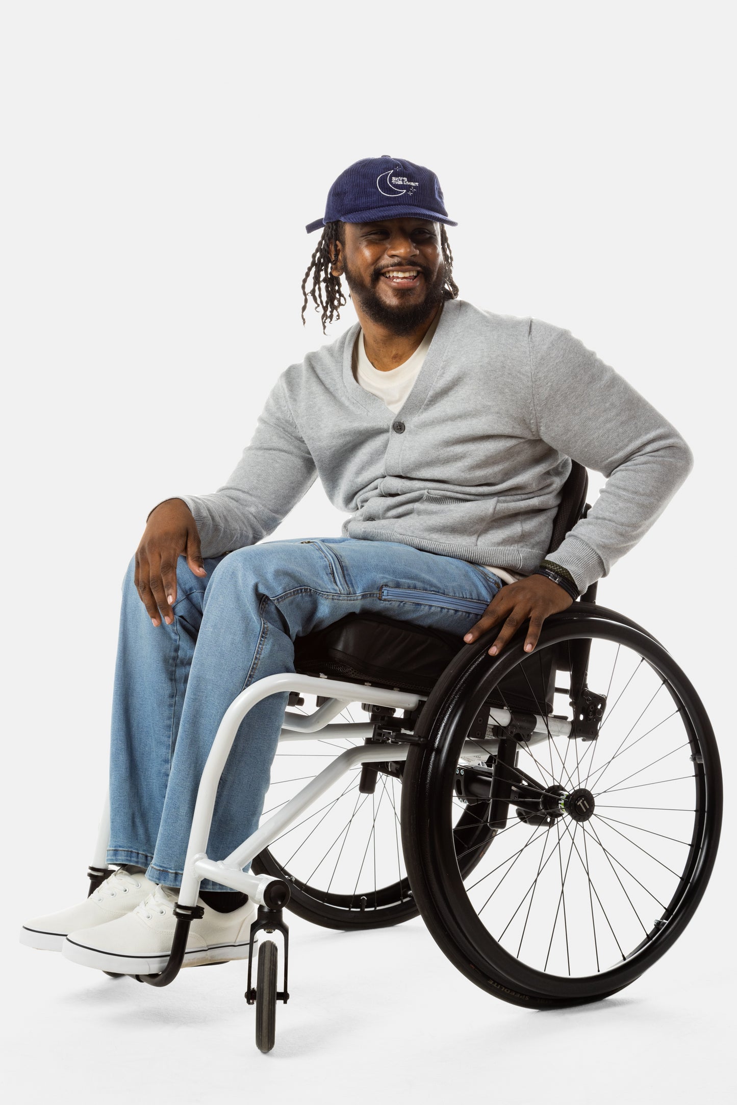 Jessi, a Black man on a white wheelchair (with short locs with a bun on the top of his hair, a goatee, cross earrings, and white sneakers) wears the No Limbits Adaptive Men's Light Wash Wheelchair Pants.