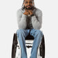 Jessi, a Black man on a white wheelchair (with short locs with a bun on the top of his hair, a goatee, cross earrings, and white sneakers) wears the No Limbits Adaptive Men's Light Wash Wheelchair Pants.
