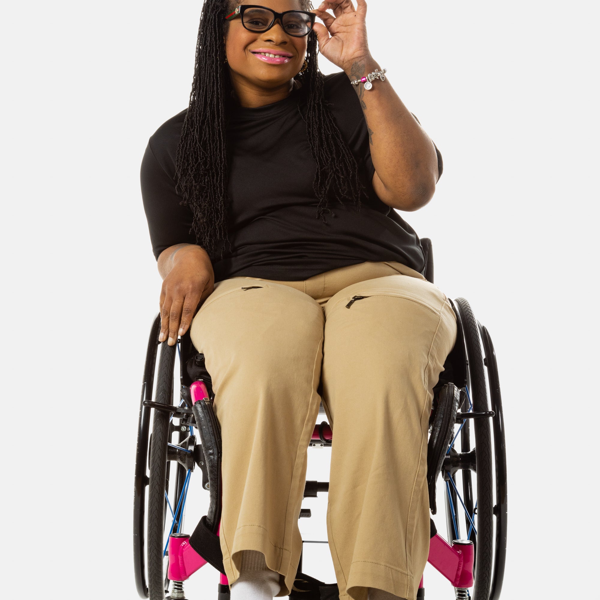 Princess, a Black woman on a pink wheelchair (with long locs, light pink lipstick, clear black sunglasses, and hoops) wears the No Limbits Adaptive Women's Khaki Wheelchair Pants. She wears sunglasses.