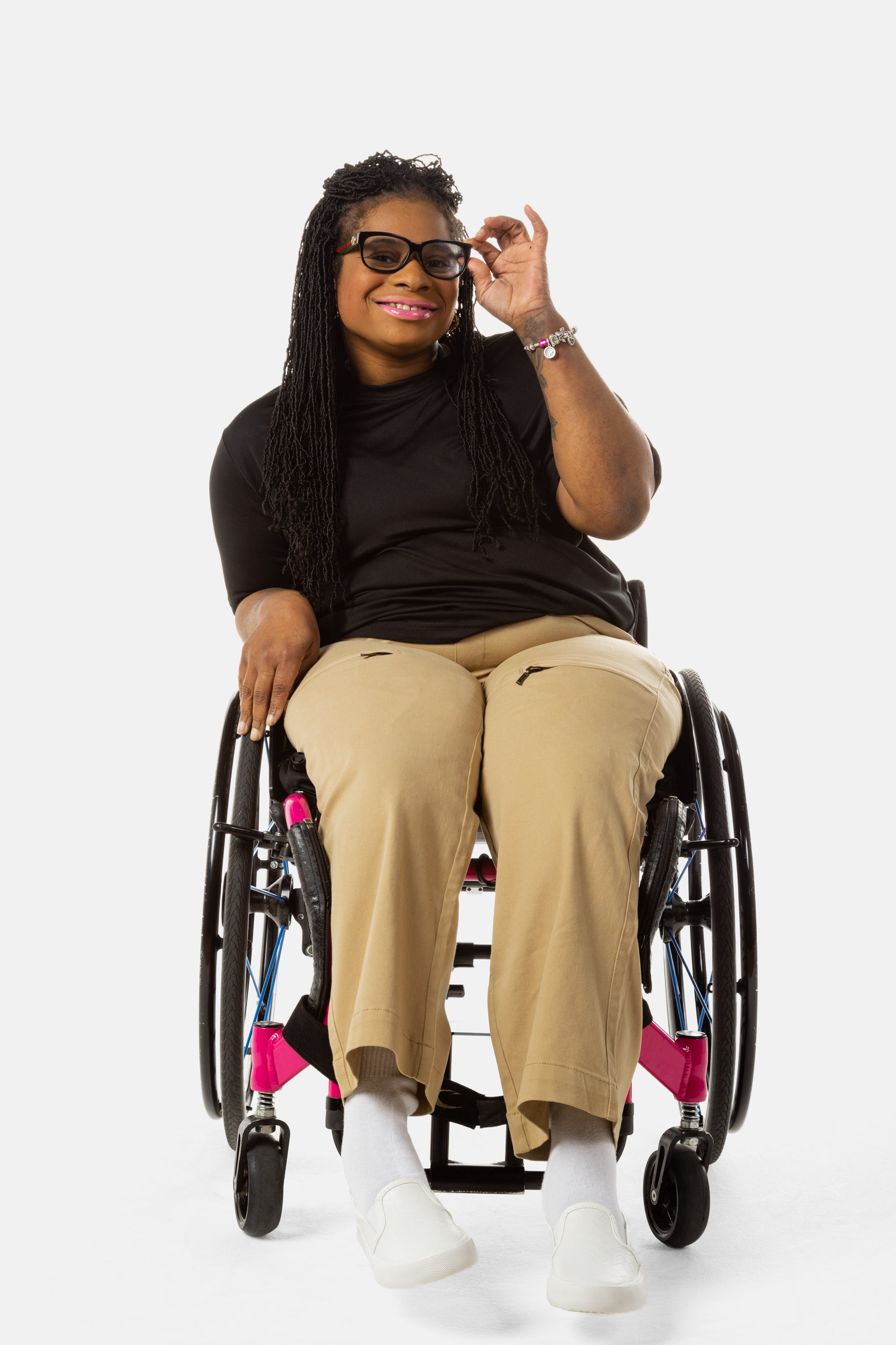 Princess, a Black woman on a pink wheelchair (with long locs, light pink lipstick, clear black sunglasses, and hoops) wears the No Limbits Adaptive Women's Khaki Wheelchair Pants. She wears sunglasses.