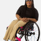 Princess, a Black woman on a pink wheelchair (with long locs, light pink lipstick, clear black sunglasses, and hoops) wears the No Limbits Adaptive Women's Khaki Wheelchair Pants.