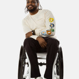 Jessi, a Black man on a white wheelchair (with short locs with a bun on the top of his hair, a goatee, cross earrings, and white sneakers) wears the No Limbits Adaptive Men's Black Wheelchair Pants.