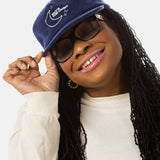 A black woman with sunglasses, pink lipstick, and long black locs wears a dark blue baseball cap that has a illustration of the moon and the stars that says in white font "Sky's the Limbit."