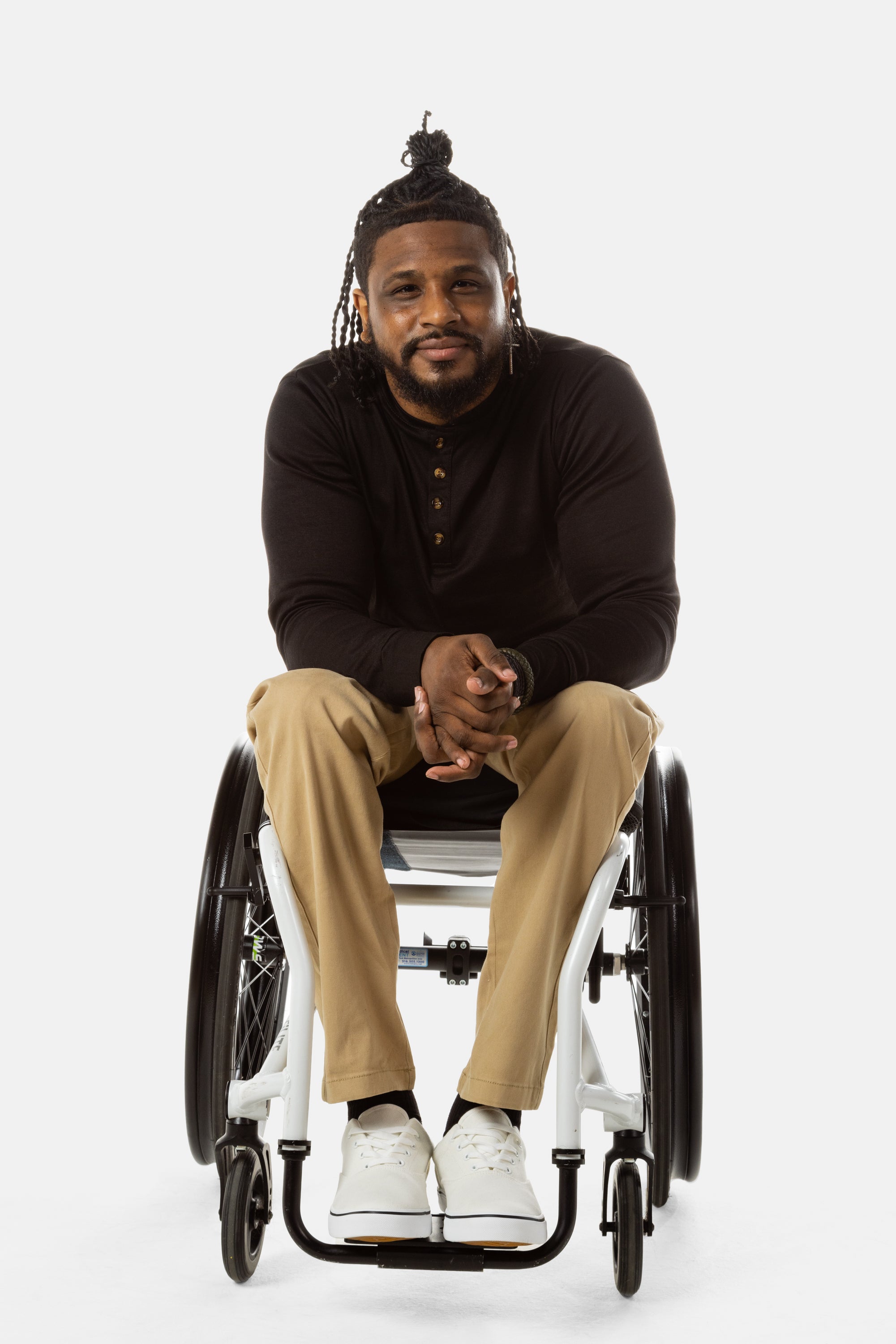 Jessi, a Black man on a white wheelchair (with short locs with a bun on the top of his hair, a goatee, cross earrings, and white sneakers) wears the No Limbits Adaptive Men's Khaki Wheelchair Pants.