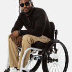 Jessi, a Black man on a white wheelchair (with short locs with a bun on the top of his hair, a goatee, cross earrings, and white sneakers) wears the No Limbits Adaptive Men's Khaki Wheelchair Pants. He is wearing black sunglasses.