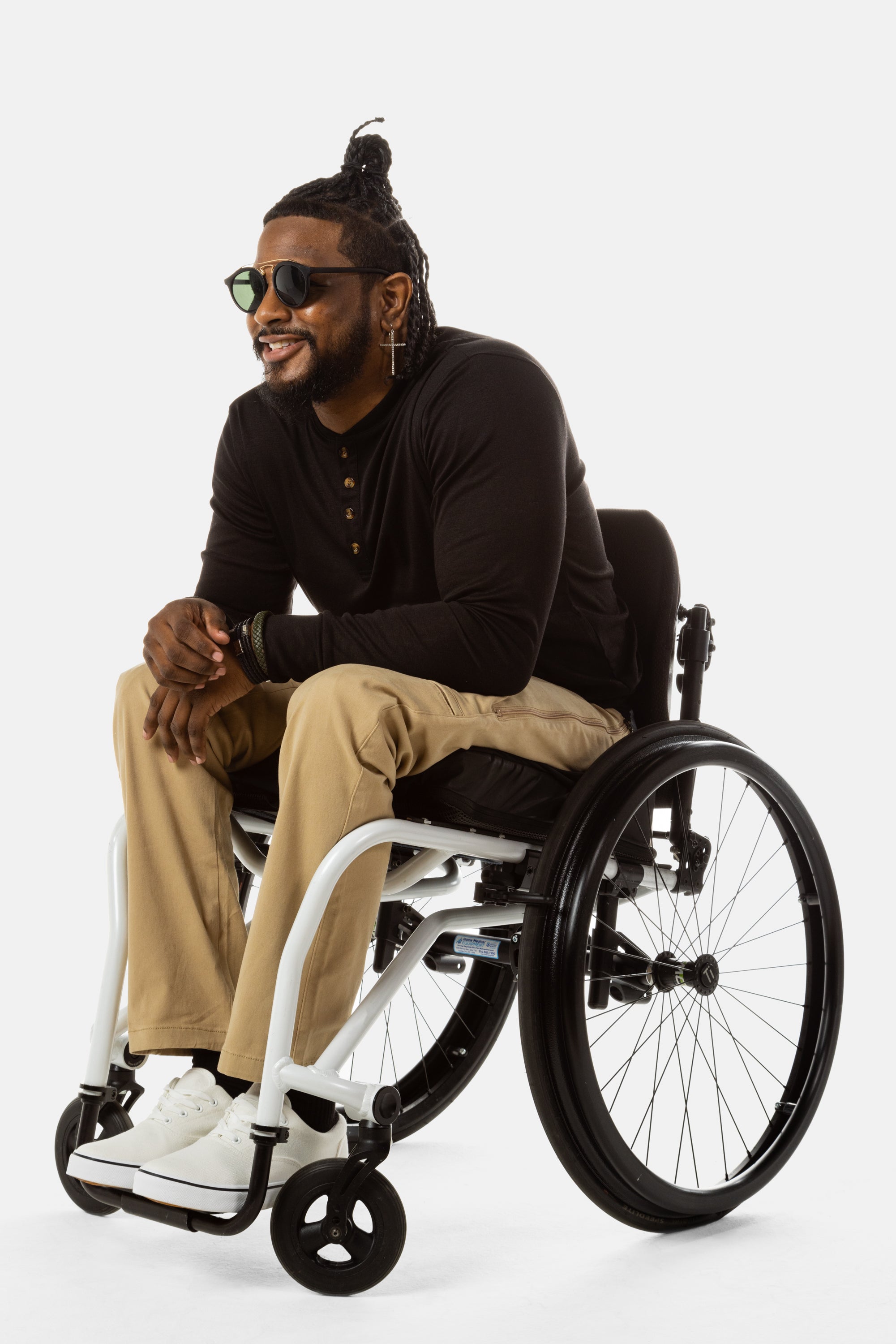 Jessi, a Black man on a white wheelchair (with short locs with a bun on the top of his hair, a goatee, cross earrings, and white sneakers) wears the No Limbits Adaptive Men's Khaki Wheelchair Pants. He is wearing black sunglasses.