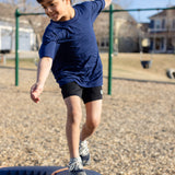 A little boy playing in the park, he is wearing a navy basic crewneck t-shirt and black shorts. You can see swing sets blurred out in the background of the photo.